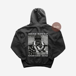 May include: Black hooded sweatshirt featuring a black and white image of ASAP Rocky with an American flag design. The text "ASAP ROCKY" and "LONG LIVE ASAP" are printed above and below the image, respectively. The sweatshirt is a casual style.