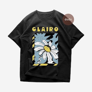May include: A black short-sleeve t-shirt featuring the text CLAIRO in yellow letters above a rectangular graphic. The graphic depicts a white daisy with a yellow and black face center against a light blue background with black shapes.