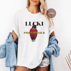 以下が含まれることがあります： 黒と金の「FREEWAVE 3」の上に「LUCKI」という文字が入った白いTシャツ。赤い髪の人のグラフィックが描かれています。柔らかい素材で作られたシャツです。