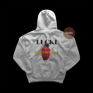 以下が含まれることがあります： 「LUCKI」の文字が黒いブロック体で、その下に金色の「FREEWAVE 3」と書かれたライトグレーのパーカー。赤いジャケットを着た人物のグラフィックが中央に配置されています。パーカーにはフードと長袖が付いています。