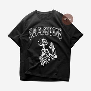 Op de afbeelding: Zwart T-shirt met de witte tekst "SUICIDEBOYS" boven een witte skeletillustratie met gevouwen handen in gebed. Het T-shirt heeft een ronde hals en korte mouwen.