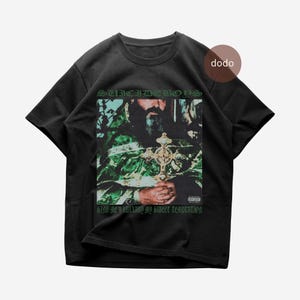 Op de afbeelding: Zwart T-shirt met een afbeelding van een man die een kruis vasthoudt, met de tekst "SUICIDEBOYS" erboven en "SING ME A LULLABY MY SWEET TEMPTATION" eronder. De afbeelding heeft een groen en bruin kleurenschema.