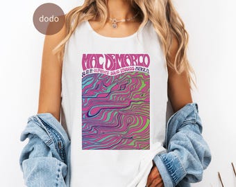 May include: White tank top featuring a graphic design with the text "Mac Demarco" in pink, with additional text and a colorful, wavy pattern in pink, blue, and green. The tank top is sleeveless.