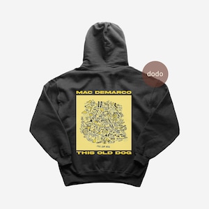 Peut inclure: Un sweat-shirt à capuche gris anthracite avec un grand carré jaune imprimé au dos. Le graphique contient des illustrations au trait noir et le texte MAC DEMARCO et THIS OLD DOG en lettres capitales jaunes au-dessus et en dessous du carré.