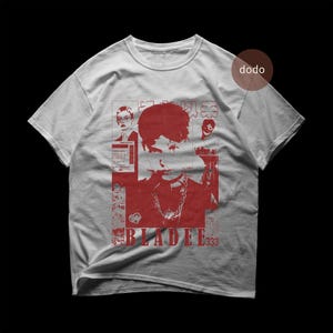 Puede incluir: Camiseta gris claro con un diseño gráfico rojo. El diseño incluye un retrato, texto y caracteres japoneses. La palabra "BLADEE" y el número "333" también son visibles.