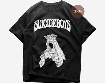 Puede incluir: Una camiseta negra de manga corta con el texto blanco SUICIDEBOYS en una fuente estilizada en la parte superior. Debajo del texto hay una ilustración gráfica blanca de una figura encapuchada con corona que sostiene una daga.