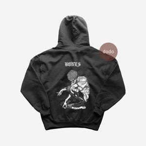 Könnte beinhalten: Schwarzer Kapuzenpullover mit einem weißen Aufdruck eines hockenden Skeletts mit Mütze. Das Wort "BONES" ist darüber gedruckt. Der Hoodie hat eine Kapuze und eine Vordertasche.