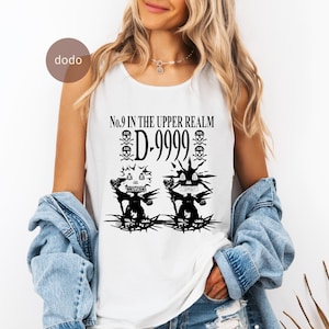 Puede incluir: Camiseta sin mangas blanca con un diseño gráfico negro. El diseño presenta el texto "No.9 IN THE UPPER REALM D-9999" y dos figuras de monstruos. El diseño incluye calaveras y elementos puntiagudos, creando una estética oscura y atrevida.