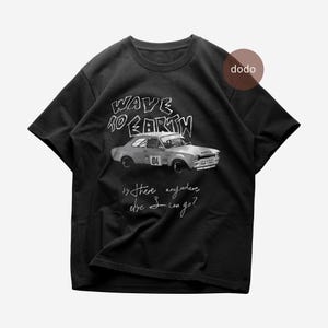 Op de afbeelding: Zwart T-shirt met een afbeelding van een oldtimer en de tekst "WAVE TO EARTH" boven de auto. Onder de auto staat de tekst "is there anywhere else I can go?"