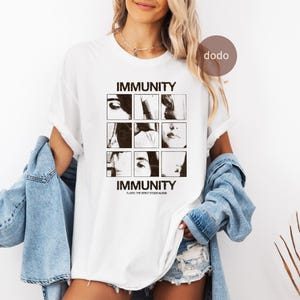 May include: A white short-sleeved t-shirt featuring a black graphic print with the text IMMUNITY at the top and bottom. The central design consists of a three-by-three grid of black and white photographic squares and the text CLAIRO: THE DEBUT STUDIO ALBUM.