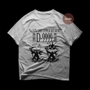 Puede incluir: Camiseta gris claro con un diseño gráfico negro. El diseño incluye el texto "No.9 IN THE UPPER REALM D-9999", calaveras, murciélagos y figuras estilizadas. La camiseta tiene cuello redondo y mangas cortas.