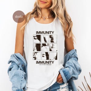 May include: A white sleeveless tank top featuring a black graphic print with the word IMMUNITY at the top and bottom. The center contains a grid of nine black and white photographic squares showing facial features, with the text CLAIRO THE DEBUT STUDIO ALBUM.