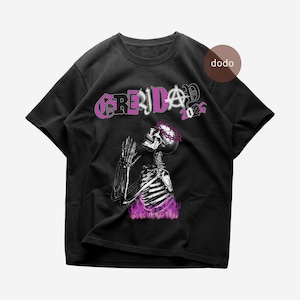 Puede incluir: Camiseta negra con un diseño de esqueleto con corona de espinas y manos rezando. El texto "GRENDA" está en morado estilizado, con "2026" en la esquina superior derecha. El esqueleto está rodeado de llamas moradas.