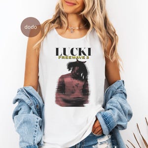 以下が含まれることがあります： 黒と緑の「LUCKI FREEWAVE 3」の文字が入った白いタンクトップ。テキストの下には、ドレッドヘアの人のぼやけた画像があります。タンクトップはノースリーブで、柔らかい素材で作られています。