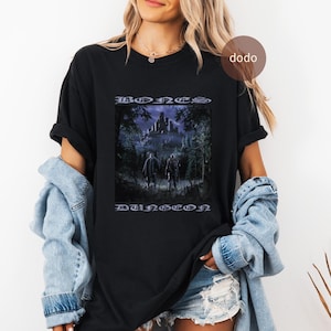 Könnte beinhalten: Schwarzes T-Shirt mit einem Fantasy-Motiv und den Worten "BONES DUNGEON". Die Grafik zeigt zwei Figuren, die auf ein Schloss in einem dunklen Wald zugehen. Das T-Shirt ist ein bequemer, lässiger Stil.