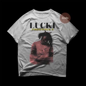 以下が含まれることがあります： 黒と黄色の「LUCKI FREEWAVE 3」の文字が入った、明るい灰色のTシャツ。赤と黒の人物のグラフィックが描かれています。Tシャツは柔らかい素材で作られています。