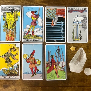 May include: Seven tarot cards arranged in two rows, including The Fool, Page of Swords, Nine of Swords, Ace of Swords, Queen of Wands, Two of Pentacles, and Six of Wands. A clear quartz crystal, a small star-shaped stone, and a round white stone.