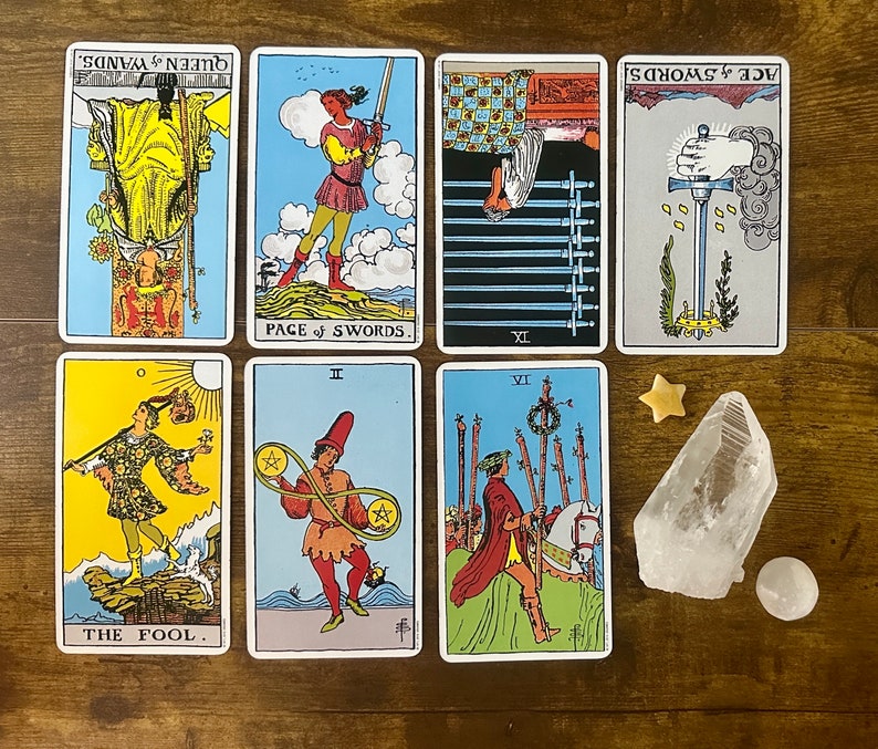 May include: Seven tarot cards arranged in two rows, including The Fool, Page of Swords, Nine of Swords, Ace of Swords, Queen of Wands, Two of Pentacles, and Six of Wands. A clear quartz crystal, a small star-shaped stone, and a round white stone.