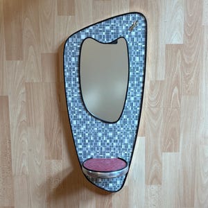 May include: A wall-mounted mirror with an asymmetrical, amoeba-like shape. The frame features a gray and white mosaic tile pattern with a black border. A small, semi-circular shelf with a red textured surface is attached at the bottom.