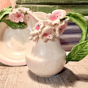 May include: A ceramic pitcher and matching dish set. The pitcher is off-white with pink floral accents and a green handle. The dish is round with a pink rim. The set is decorated with pink flowers and green leaves.