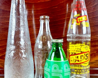 May include: Four glass bottles, two clear and two green, with various labels and text. The bottles are on a wooden surface. The text on the bottles includes "Dad's", "Original Jamaica Ginger", "7 fl oz", "Return this bottle for refund", and "Net 16 fl oz (1 pt)."