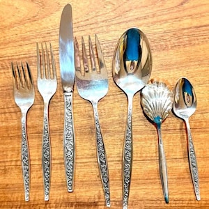 May include: A collection of vintage silverware, including forks, knives, and spoons, arranged on a wooden surface. The silverware has ornate, engraved handles and a shiny, reflective finish. The set includes a serving spoon and a decorative spoon.
