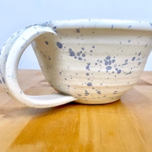 May include: A white ceramic bowl with a speckled blue glaze and a unique handle. The bowl is sitting on a wooden surface.