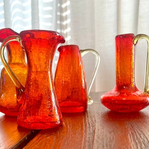 May include: Five small, red glass pitchers with textured surfaces and yellow handles. The pitchers are arranged on a wooden table.