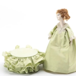 Könnte beinhalten: Eine Porzellanpuppe in einem hellgrünen Kleid mit Spitzenbesatz steht auf einem runden, mehrstufigen, hellgrünen Sockel.