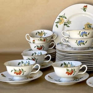 May include: A set of white porcelain teacups and saucers with floral designs in shades of blue, yellow, red, and green. The cups have a handle on one side and a small foot on the bottom. The saucers have a scalloped edge.