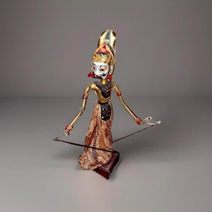 May include: A traditional Indonesian Wayang Golek puppet. The puppet has a gold and red headdress, a white face with red accents, and a gold and brown patterned costume. The puppet is standing on a small, red base and holding two thin rods.