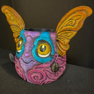 May include: A whimsical, colorful, 3D-printed creature-shaped container. The container features large, ornate wings in shades of orange and yellow, with a face in purple, blue, and pink. The eyes are yellow with black pupils, and the container has a black interior.
