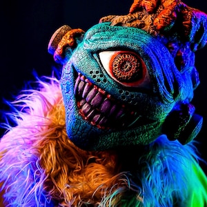 May include: A close-up of a fantastical creature mask with a textured, blue-green face, large red eye, and sharp purple teeth. The mask has orange and brown details on the head and is surrounded by a fluffy, multicolored collar.
