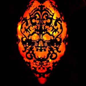Puede incluir: Una calabaza naranja iluminada, tallada con una calavera y un elaborado diseño de filigrana negra. El contraste entre el negro y el naranja crea un ambiente festivo, perfecto para decoraciones de Halloween.