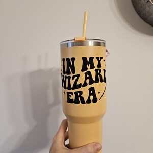 Puede incluir: Un vaso de acero inoxidable amarillo con una calcomanía de vinilo negro que dice "In My Wizard Era".