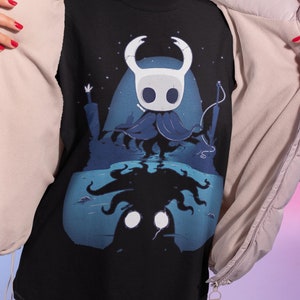 May include: Black t-shirt with a blue and white illustration of a character from the video game Hollow Knight. The character is standing in front of a dark blue background with a starry sky. The character's reflection is visible in the water below.