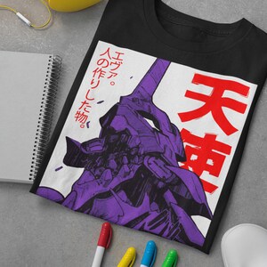 Może przedstawiać: Czarny T-shirt z białym nadrukiem przedstawiającym fioletowego robota z japońskim tekstem "エヴァ。 人の作りし物。 天  " w kolorze czerwonym.