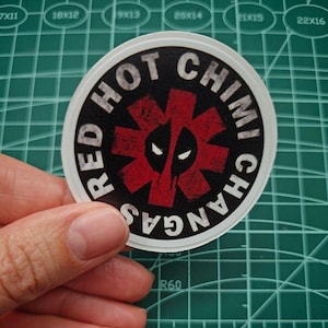 May include: A round sticker with a red and black design. The sticker features a red star with a black outline and a black skull with white eyes. The text "RED HOT CHILI PEPPERS" is written around the star in a white circle.