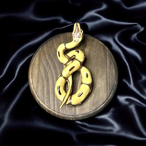 Pode incluir: Escultura de cobra amarela e castanha sobre uma base redonda de madeira. A cobra está enrolada e tem marcas castanhas escuras. A base de madeira é de cor castanha escura. O fundo é um tecido escuro e sedoso.