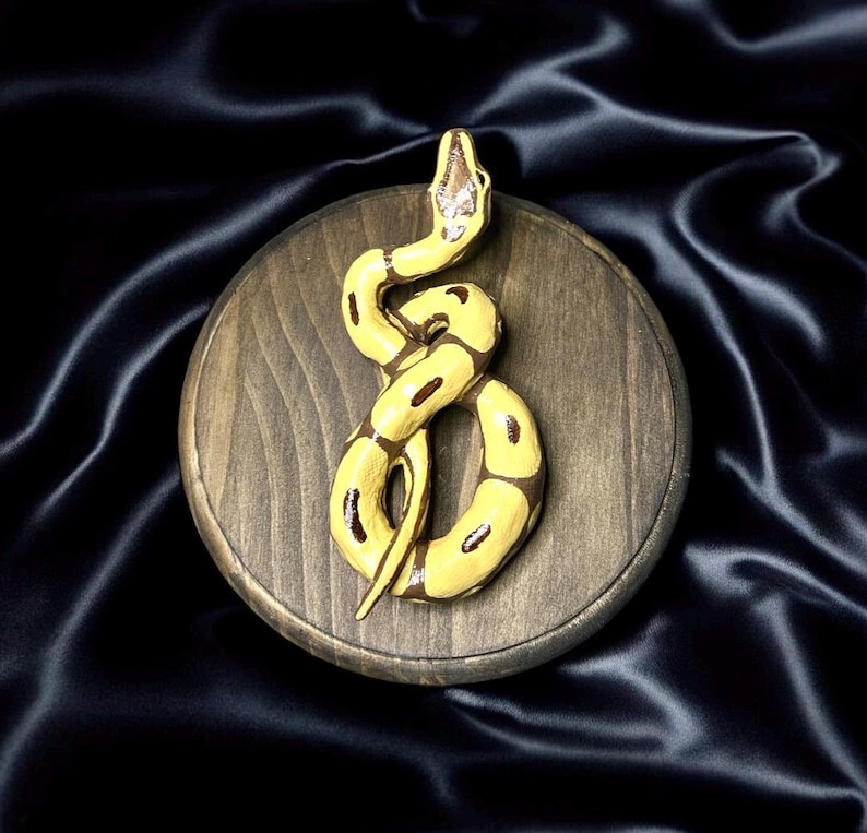 May include: A yellow and brown snake sculpture on a round, wooden base. The snake is coiled and has dark brown markings. The wooden base is a dark brown color. The background is a dark, silky fabric.