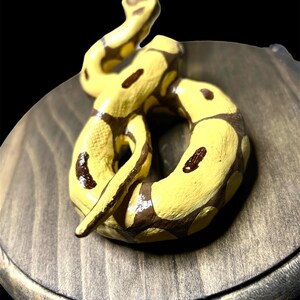 May include: A hand-painted ceramic snake figurine in yellow and brown, coiled on a wooden base. The snake has dark brown spots and is posed in a dynamic, curved shape. The wooden base has a dark, stained finish.