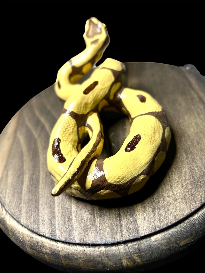 May include: A hand-painted ceramic snake figurine in yellow and brown, coiled on a wooden base. The snake has dark brown spots and is posed in a dynamic, curved shape. The wooden base has a dark, stained finish.