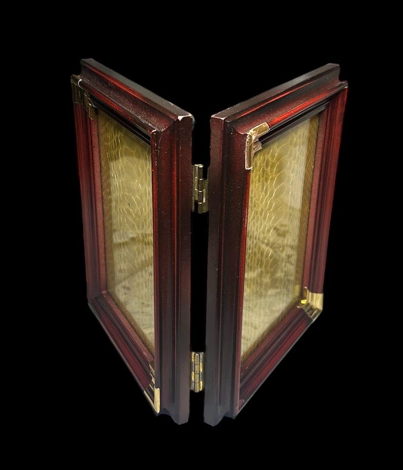 May include: A two-panel, hinged picture frame with a dark red wooden frame and gold-coloured corner accents. The frame is open, revealing a textured, golden-coloured interior. The hinges are gold-coloured.