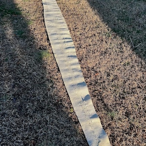 Puede incluir: Una larga tira de tela beige texturizada descansa sobre hierba seca. La tira tiene un patr&oacute;n repetitivo y est&aacute; ligeramente curvada. La tela parece estar hecha de un material tejido. La imagen est&aacute; tomada al aire libre con luz natural.