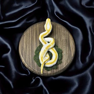 May include: A decorative sculpture of a yellow and white snake coiled on a round wooden plaque. The snake rests on a bed of green moss. The background is a dark, silky fabric.