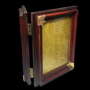 May include: A hinged, claret-coloured picture frame with gold corner detailing. The frame contains a textured, yellow-green material, possibly leather or a similar pattern. The frame is open, revealing the hinge and the material inside.