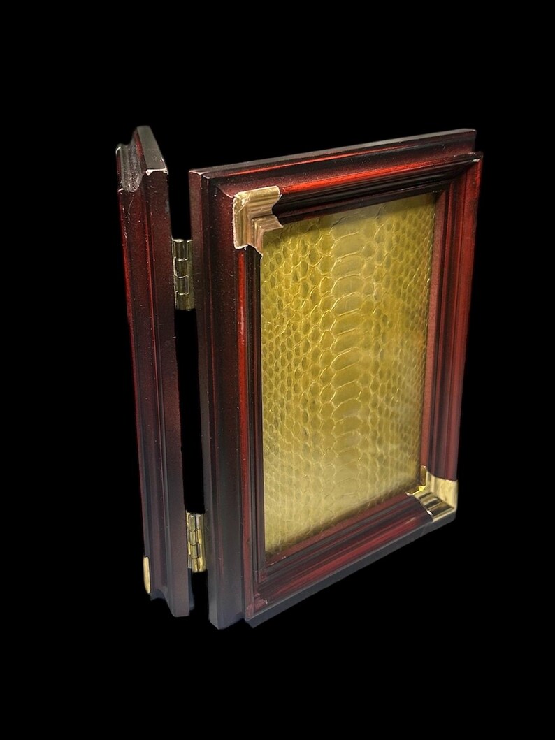 May include: A hinged, claret-coloured picture frame with gold corner detailing. The frame contains a textured, yellow-green material, possibly leather or a similar pattern. The frame is open, revealing the hinge and the material inside.
