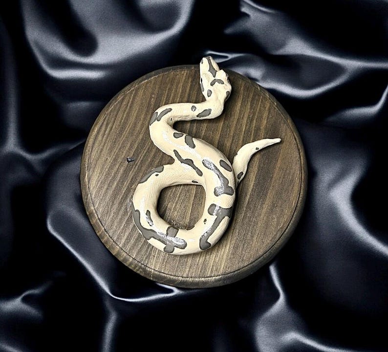 K&ouml;nnte beinhalten: Eine cremefarbene Schlangenskulptur mit grauen Markierungen, auf einer runden Holzbasis gewunden. Die Schlange hat eine strukturierte Oberfl&auml;che. Die Holzbasis hat eine nat&uuml;rliche Maserung. Der Hintergrund ist ein dunkler, seidiger Stoff.