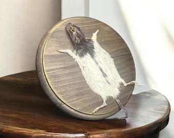 May include: A taxidermied rat mounted on a round wooden plaque. The rat has black and white fur and is posed with its limbs outstretched. The plaque has a gold-painted edge and a wood grain finish.