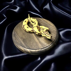 May include: A detailed, handcrafted figurine of a yellow and brown snake coiled on a round, wooden base. The snake's scales are meticulously painted, and the base has a dark, weathered finish. The background is a dark, silky fabric.